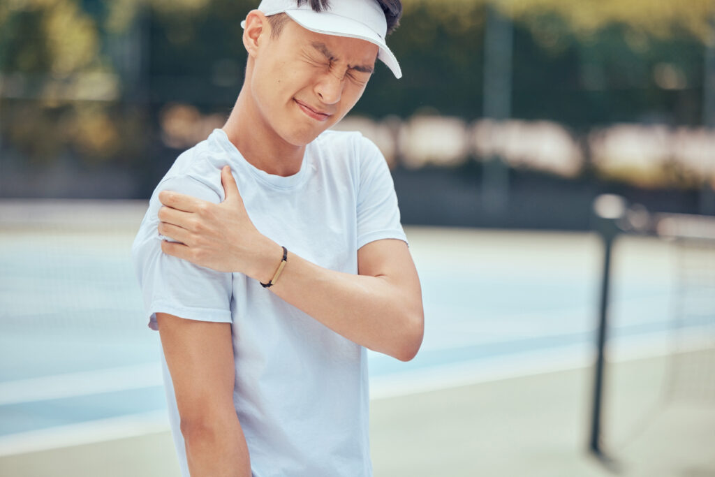 Biological Rotator Cuff Repair With Tetrous EnFix, CONMED, And Arthrex BioSurge® Products Young man in a white shirt and visor rubs his shoulder with a pained expression on a tennis court, wincing in pain.