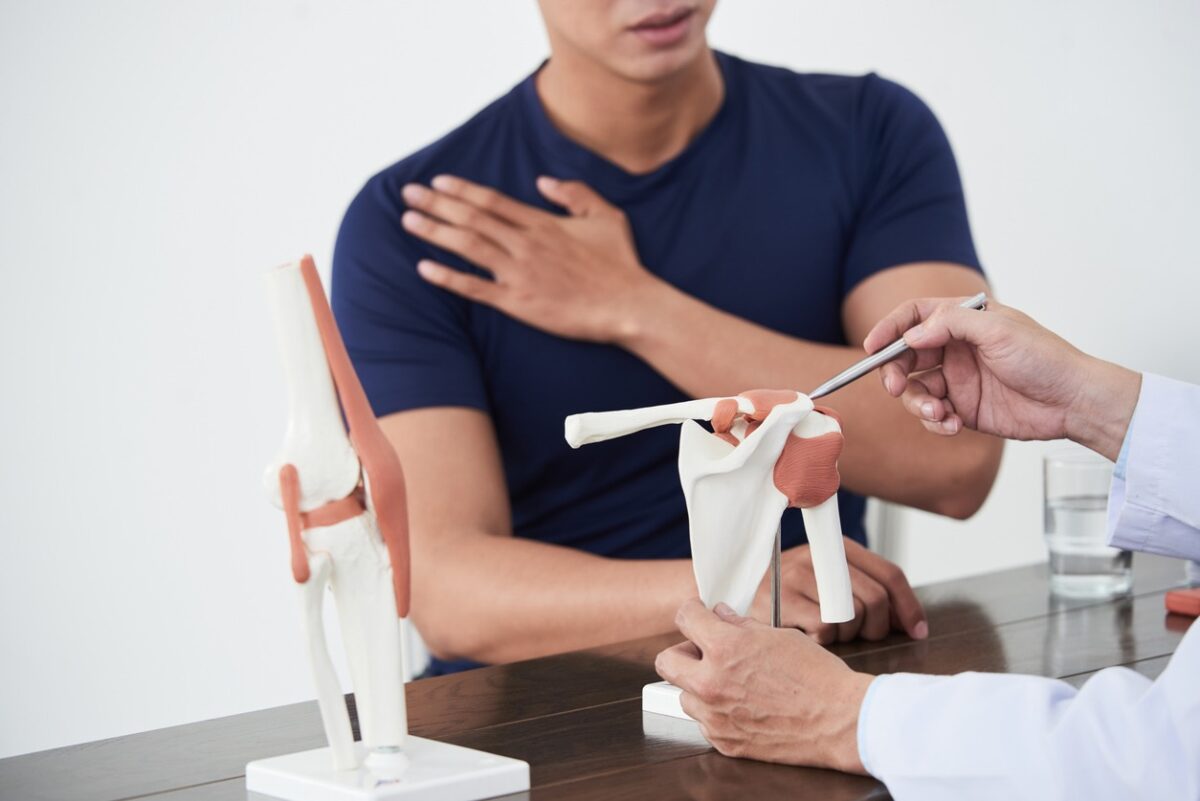 Doctor demonstrates a knee joint model to a patient, pointing with a tool as the patient places a hand on his chest during a consultation.