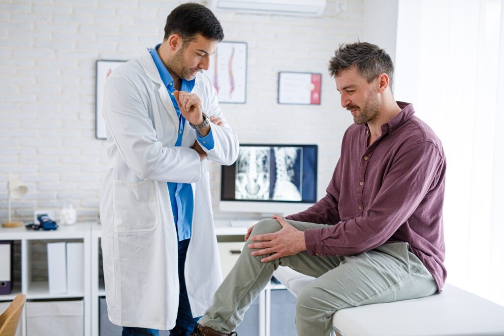 Doctor in a white coat consults with a seated patient, who is holding his knee during a clinic visit with X-ray images on a monitor in the background.