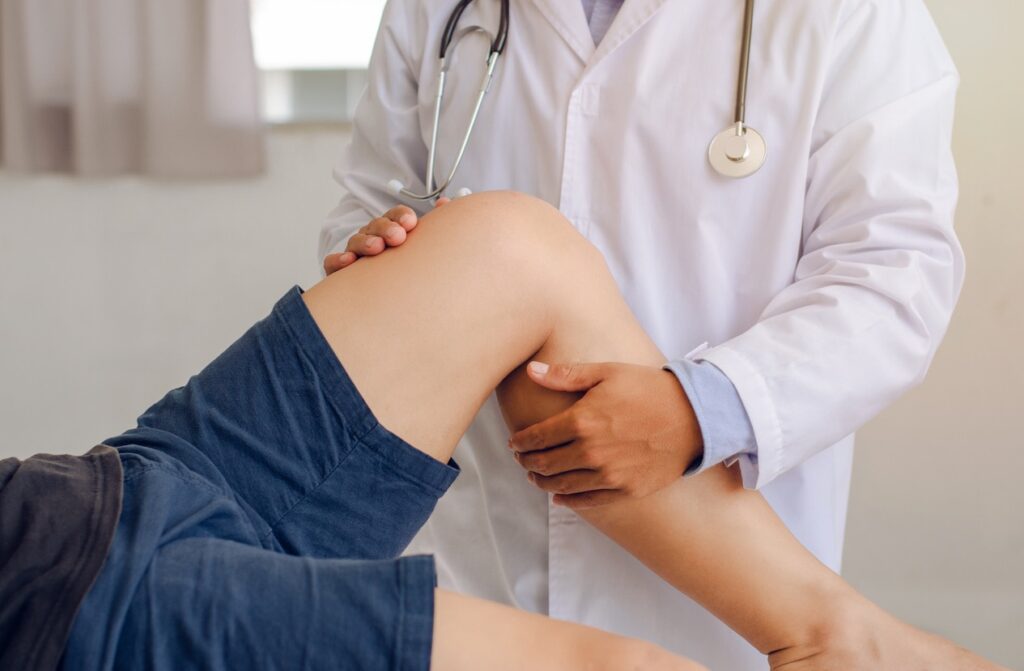 Physician in a white coat with stethoscope stabilizing and examining a patient&rsquo;s bent knee during a medical check-up.