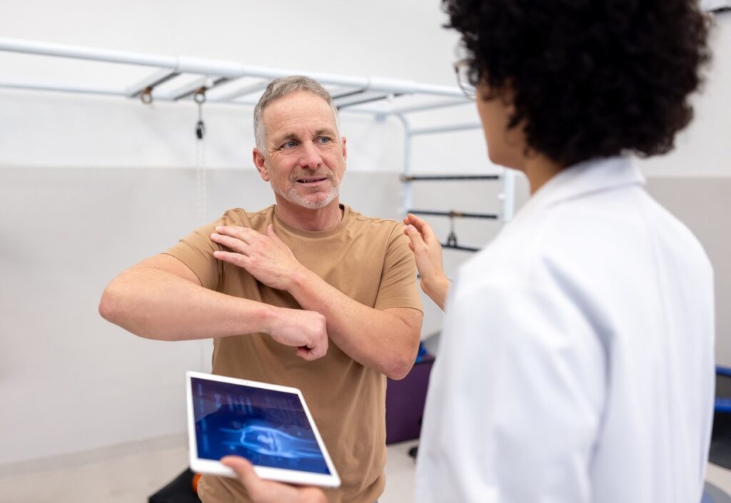 Middle-aged man stretches his shoulder while a clinician watches and holds a tablet showing a blue image in a clinical setting.