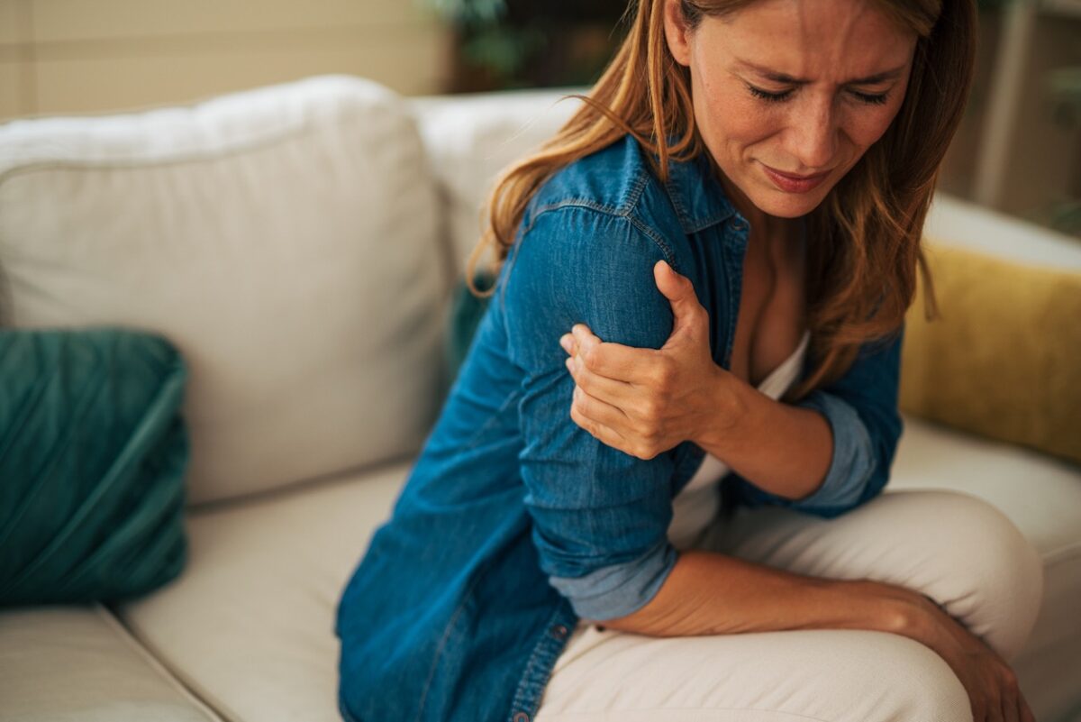 Woman on a beige sofa grimacing and rubbing her shoulder in discomfort.