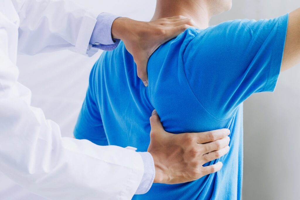 Medical professional examining a patient's upper back and shoulder for mobility or pain assessment; clinical setting shows hands on shoulder area.