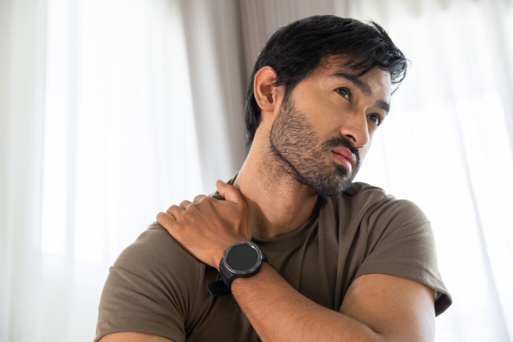 Man wearing a brown t-shirt touches his left shoulder with his right hand, looking off to the side in a softly lit room.