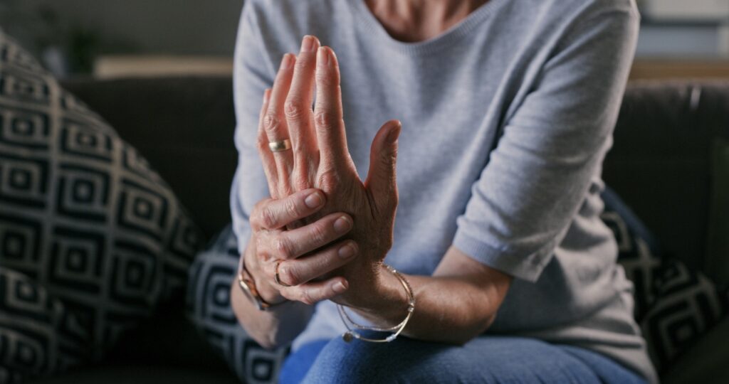 Close-up of hands clasped in prayer, wearing rings and bracelets, resting on a knee on a sofa.